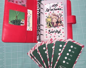 Peut inclure: Agenda en cuir rouge avec un classeur argenté, ouvert pour révéler des inserts d'épargne Grinchmas. Comprend des cartes à rayures vertes et blanches avec des montants d'épargne et un total de 160 €. Un charm de sapin de Noël et une carte "Naughty" sont également visibles.