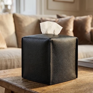 Puede incluir: Un dispensador de pañuelos cuadrado negro hecho de material texturizado. Un pañuelo blanco sobresale de la parte superior. El dispensador está sobre una mesa de madera, con un sofá al fondo.
