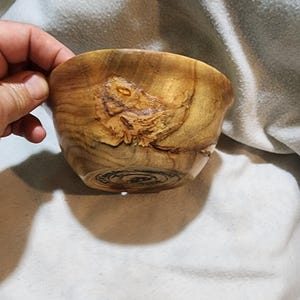 Puede incluir: Un cuenco de madera hecho a mano con un acabado marrón claro natural. El cuenco presenta patrones de vetas de madera únicos y un diseño decorativo en la base. Una mano sostiene el cuenco, mostrando su tamaño y forma.