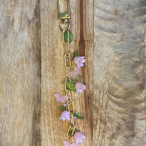 May include: A gold-colored keychain with a chain of links, adorned with pink flowers, butterflies, and green leaves. The keychain has a clasp at the top for easy attachment. The floral and butterfly design adds a touch of whimsy.
