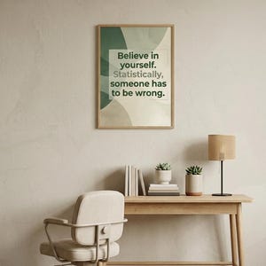 Op de afbeelding: Een ingelijste kunstprint met abstracte groene en beige organische vormen met de tekst: Believe in yourself. Statistically, someone has to be wrong. De print is ingelijst in een licht houten frame en hangt aan een neutrale muur.