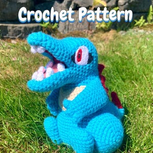 May include: A blue crochet creature with a cream-colored belly, red back spikes, and an open mouth showing white teeth. The eyes are red with black outlines. The text "Crochet Pattern" is displayed in white letters with a blue outline above the toy.