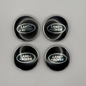May include: Four black Land Rover wheel center caps with a silver trim. Each cap features the Land Rover logo in white and green. The caps are arranged in a square formation against a light gray background.