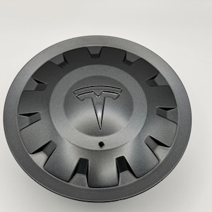 May include: A dark gray wheel center cap with the Tesla logo in the center. The cap has a circular design with angular cutouts around the edge. The Tesla logo is a stylized "T" in silver.