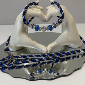 May include: A braided dog collar and lead set in blue, brown, and silver. The collar has blue and silver gemstone embellishments. The lead has a silver clasp. The set is presented on a reflective surface.