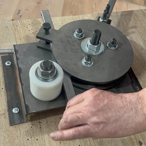 May include: A metal bending tool with a circular plate, a white roller, and a lever. The tool is bolted to a rectangular metal base and is designed for shaping metal. The image shows the tool in use.
