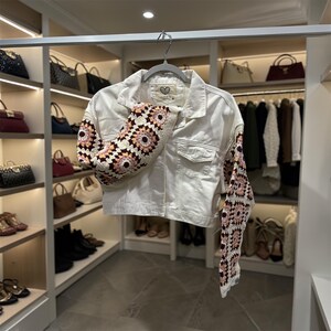 May include: A white denim jacket with crochet sleeves. The sleeves feature a floral pattern in shades of pink, cream, and brown. The jacket has a classic collar, button-up front, and two chest pockets. The jacket is hanging on a gray hanger.