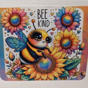 May include: A colorful, rectangular design featuring a cartoon bee with large eyes, wings, and a striped body, surrounded by sunflowers. The text "BEE KIND" is displayed above the bee. The design includes hearts and a rainbow color scheme.
