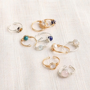 Peut inclure: Plusieurs bagues faites à la main, enveloppées de fil métallique. Les bagues présentent diverses pierres précieuses, dont des perles, de la turquoise et du quartz rose. Le fil est argenté et doré. Les bagues sont présentées sur un tissu crème.