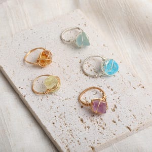 Peut inclure: Cinq bagues en pierres précieuses avec des montures en fil de fer. Les bagues présentent des pierres de différentes couleurs, notamment orange, jaune, bleu et violet. Les anneaux sont de couleur or ou argent. Les bagues sont présentées sur une surface texturée de couleur claire.
