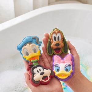 May include: Three colorful bath bombs shaped like cartoon character faces. One features a blue-hatted duck, another a brown dog with green ears, and the third a white duck with a pink bow. The fourth is a black and white dog with a red base.
