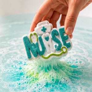 May include: A bath bomb in the shape of the word "NURSE" with a nurse's hat and stethoscope design. The bath bomb is white with teal and green accents and is dissolving in blue water, creating bubbles.