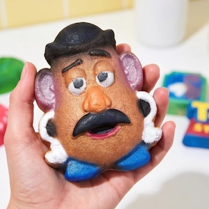 Könnte beinhalten: Eine Badebombe in Form von Mr. Potato Head mit einem braunen, kartoffelartigen Körper, einem schwarzen Bowlerhut, einem schwarzen Schnurrbart, weißen Ohren und blauen Schuhen. Das Gesicht ist mit Augen, Brauen und einem roten Mund bemalt.