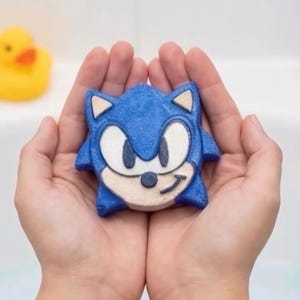 以下が含まれることがあります： ソニック・ザ・ヘッジホッグの形をした青いバスボム。白とベージュの顔の特徴があります。バスボムは両手で包み込まれ、背景には黄色のラバーダックがいます。