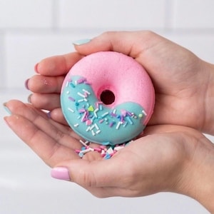 Puede incluir: Una bomba de baño en forma de donut rosa y azul con chispas de colores. La bomba de baño se sostiene en manos abiertas, mostrando sus colores vibrantes y su diseño lúdico. El donut es mitad rosa y mitad azul, con chispas en el lado azul.