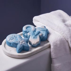 Könnte beinhalten: Eine Sammlung von blau-weißen geformten Seifen in Form eines Seesterns, eines Babybodys, eines Babyfläschchens und einer runden Scheibe mit Sanddollar-Muster, angeordnet auf einem ovalen weißen Marmortablett neben einem gefalteten weißen Handtuch.
