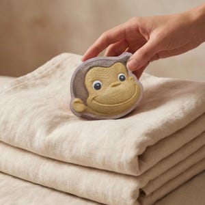 Könnte beinhalten: Ein affenförmiger Badeschaum in Beige-, Braun- und Gelbtönen mit einem lächelnden Gesicht. Er liegt auf einem Stapel gefalteter, hellfarbiger Stoffe. Das Bild ist gut beleuchtet und zeigt die Details des Badeschaums und die Textur des Stoffes.