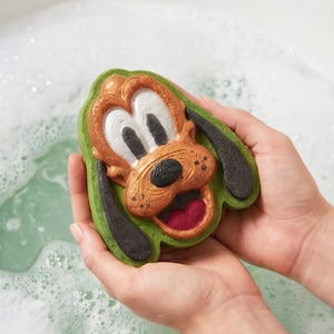 Puede incluir: Una bomba de baño con forma de cara de perro de dibujos animados en colores bronceado, blanco, negro y rojo. El personaje presenta grandes ojos blancos con pupilas negras, una nariz negra y una boca abierta que muestra una lengua roja.