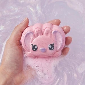 Puede incluir: Una bomba de baño rosa con forma de animal de dibujos animados con ojos y orejas grandes. La bomba de baño se sostiene en una mano y está rodeada de agua rosa y burbujeante. La bomba de baño tiene una superficie texturizada.