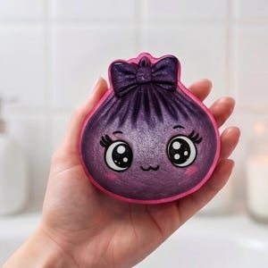 May include: A round, purple, shimmering bath bomb shaped like a dumpling with a dark purple bow on top. The surface features a printed face with large black and white eyes, long eyelashes, a small mouth, and pink blush marks on the cheeks.