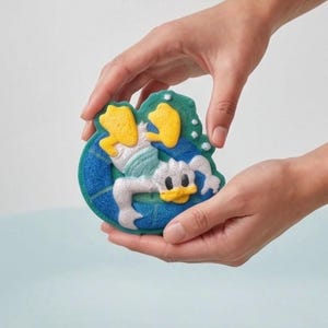 May include: A bath bomb shaped like Donald Duck, featuring yellow, white, and blue colors. The bath bomb is held in two hands, with a light blue background.
