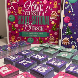 Puede incluir: Una caja de calendario de adviento con fondo rosa brillante que presenta iconos festivos como renos, casas de jengibre y bastones de caramelo, con el texto Have Yourself A Scent-Sational Season, junto a pilas de tarjetas cuadradas numeradas.