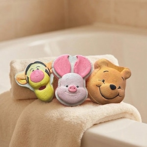 May include: Three character-shaped bath toys: Tigger, Piglet, and Winnie the Pooh, on a folded beige towel. Tigger is yellow and orange, Piglet is pink, and Winnie the Pooh is tan. The toys are near a white bathtub.