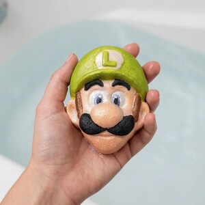 May include: A novelty bath bomb in the shape of a cartoon character's face, featuring a green hat with a white 'L', a black mustache, and brown hair. The bath bomb is held in a hand, with a light blue background.