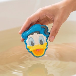 Puede incluir: Una bomba de baño moldeada con la forma de la cabeza del Pato Donald, con un sombrero de marinero azul, cara blanca, pico amarillo y ojos azules. El diseño del personaje incluye una pequeña lengua rosa visible en el pico abierto y texturizado.