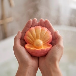 Puede incluir: Una bomba de baño con forma de concha marina con un degradado de tonos naranja y rosa. El centro de la concha presenta una esfera redonda similar a una perla. La superficie de la bomba de baño tiene una textura fina y brillante, sostenida en dos manos.