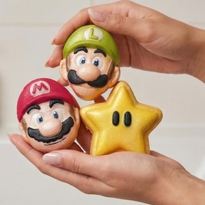 May include: Three bath bombs in the shape of Mario, Luigi, and a yellow star. The Mario bath bomb has a red hat with a white "M". The Luigi bath bomb has a green hat with a white "L". The star has two black ovals.