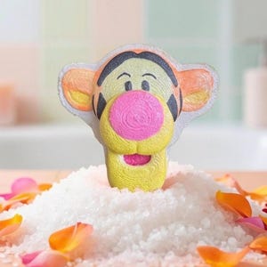 May include: A yellow bath bomb shaped like Tigger from Winnie the Pooh, with a pink nose and mouth, black stripes, and orange ears. The bath bomb is surrounded by white bath salts and colorful flower petals.