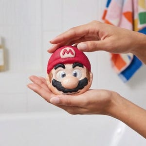 May include: A bath bomb shaped like the character Mario from the Super Mario Bros. video game series. The bath bomb features a red cap with a white "M", a black mustache, and a tan face. The bath bomb is held in open hands.