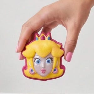May include: A Princess Peach-shaped bath bomb with yellow hair, a pink crown, and a pink outline. The bath bomb has blue eyes, pink lips, and pink earrings. The bath bomb is held by a hand with pink nail polish.