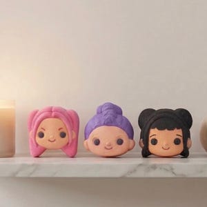 Könnte beinhalten: Drei dekorative Badebomben in Form von Cartoon-Gesichtern mit unterschiedlichen Frisuren. Die Badebomben sind rosa, lila und schwarz und sind auf einem weißen Regal angeordnet. Jedes Gesicht hat einen lächelnden Ausdruck und dunkle Augen.