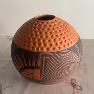 May include: A spherical terracotta vase featuring a textured upper section with carved scale-like patterns and a smooth lower section decorated with intricate black geometric line work, including sunburst motifs and triangular shapes.