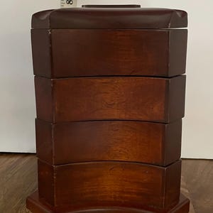 May include: A dark brown wooden storage box with four stacked, curved-front drawers. The unit stands approximately 8 inches tall as indicated by the visible ruler. The box features a smooth, polished finish and a rectangular base with a matching top.