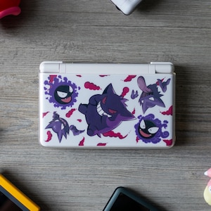 May include: A white handheld gaming console featuring a graphic print of purple Gengar and Gastly characters surrounded by red smoke patterns. The device is closed and rectangular with a hinge along the top edge, displaying a smooth, glossy finish.