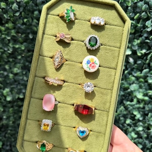 May include: A collection of sixteen assorted rings displayed on a green velvet organizer. The rings feature various designs including green marquise stones, clear crystals, a floral ceramic cabochon, pink cabochons, and colored faceted gems.