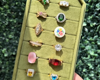 May include: A collection of sixteen assorted rings displayed on a green velvet organizer. The rings feature various designs including green marquise stones, clear crystals, a floral ceramic cabochon, pink cabochons, and colored faceted gems.