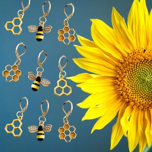 Puede incluir: Pendientes dorados con diseños de abeja y panal. Algunos pendientes presentan una abeja con rayas negras y amarillas y alas de pedrería. Un gran girasol amarillo está en el fondo sobre un fondo azul.