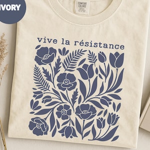 Può includere: T-shirt color avorio con un design floreale blu navy e la scritta "vive la résistance". Il design include vari fiori e foglie. La maglietta è di Comfort Colors.