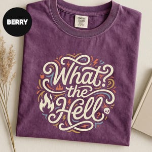 May include: Berry-colored t-shirt with the phrase "What the Hell" in a circular design. The design includes flames, lightning bolts, and a skull. The shirt is made of a soft material and has a crew neck.