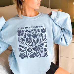 Könnte beinhalten: Hellblaues Sweatshirt mit dem Schriftzug "vive la résistance" über einem dunkelblauen Blumenmuster. Das florale Design umfasst verschiedene Blumen und Blätter. Das Sweatshirt hat lange Ärmel und einen Rundhalsausschnitt.