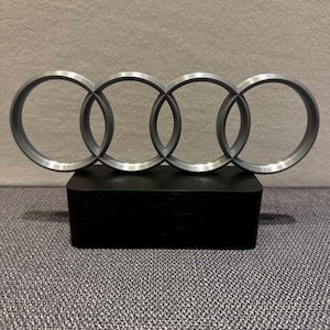 Könnte beinhalten: Ein silbernes Audi-Logo, bestehend aus vier ineinandergreifenden Ringen, wird auf einem schwarzen rechteckigen Sockel präsentiert. Das Logo ist mittig platziert und hebt sich von einem neutralen Hintergrund ab, was ein modernes Design zeigt.