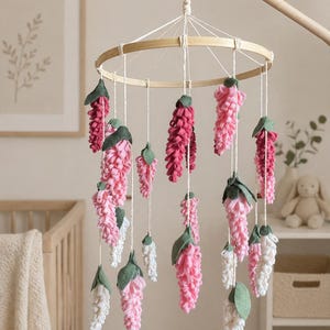 Puede incluir: Móvil para bebé con flores de fieltro en tonos rosa, blanco y burdeos, suspendidas de un aro de bambú. Las flores tienen hojas verdes y cuelgan de cuerdas blancas, creando una delicada decoración.