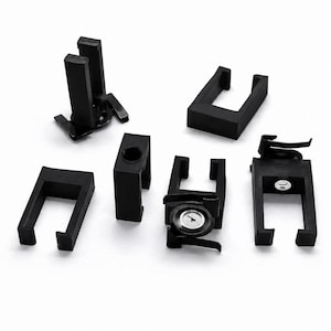 May include: A collection of six black plastic components featuring U-shaped brackets and cylindrical inserts. Some pieces include small metallic circular fasteners or screws embedded within the centre of the black plastic structures.