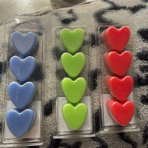 May include: Three packs of heart-shaped wax melts in blue, green, and red. Each pack contains four heart-shaped melts in a clear plastic container. The melts are arranged in a vertical row.