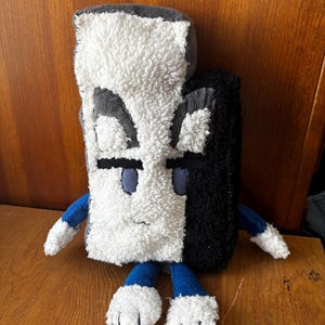 May include: A plush toy in a rectangular shape, featuring a split design of white, gray, and black. It has blue eyes, arms, and feet, with white paws. The toy is made of soft, textured fabric.