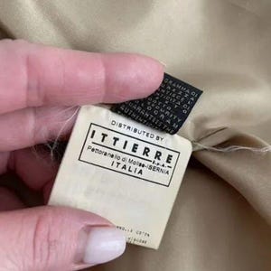 May include: A beige fabric garment interior featuring a white rectangular clothing label with black text that reads DISTRIBUTED BY ITTIERRE S.P.A. Pettoranello di Molise-ISERNIA ITALIA, positioned above a smaller black security tag.